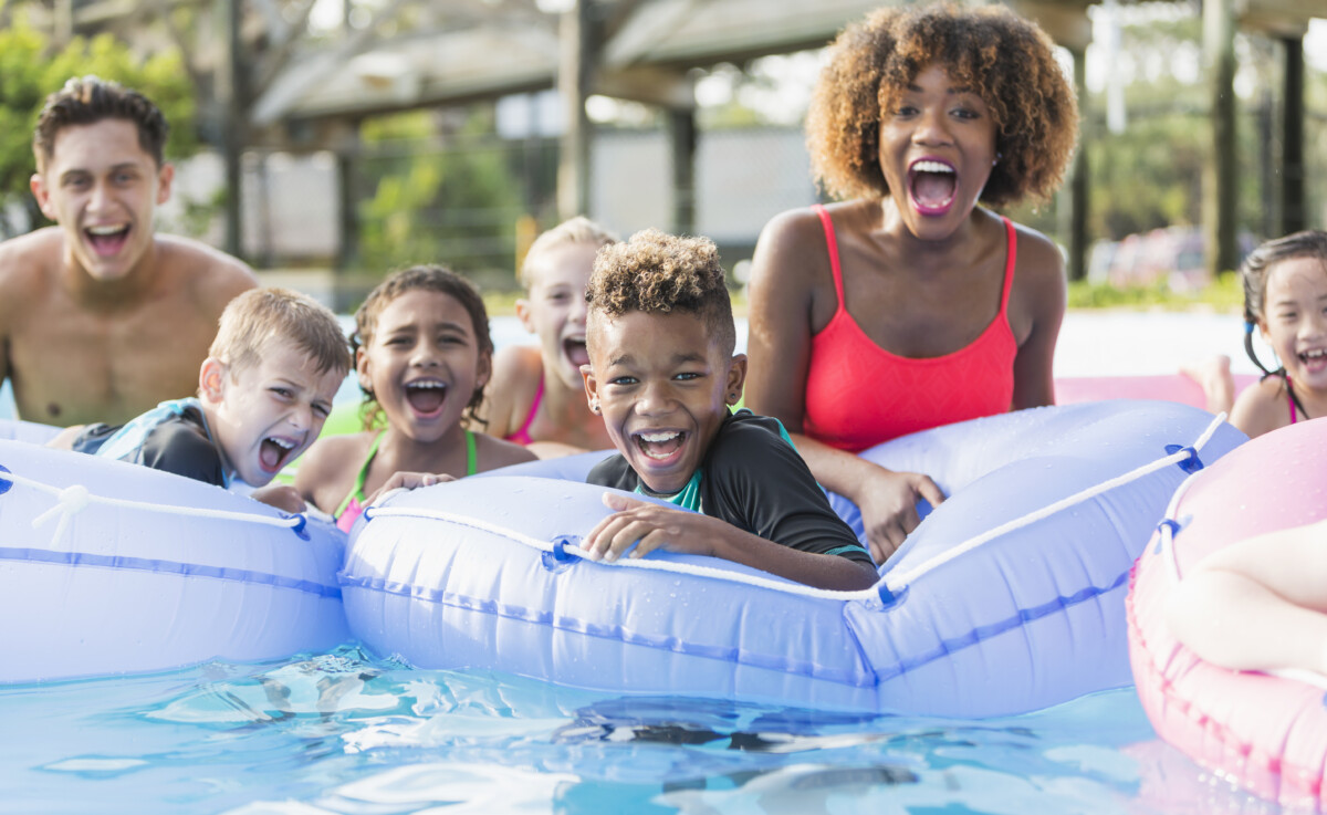 A group of multi-ethnic children and young adults on the lazy river at a water park, shouting with joy and looking at the camera. The focus is on the mixed race African American and Caucasian 9 year old boy in the center foreground.