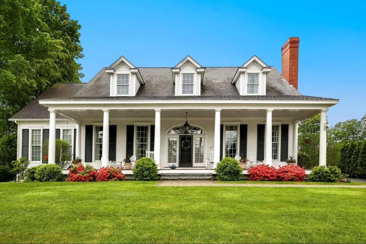 This cape cod house is white with black shutters and a central chimney includes pillars and a front porch.