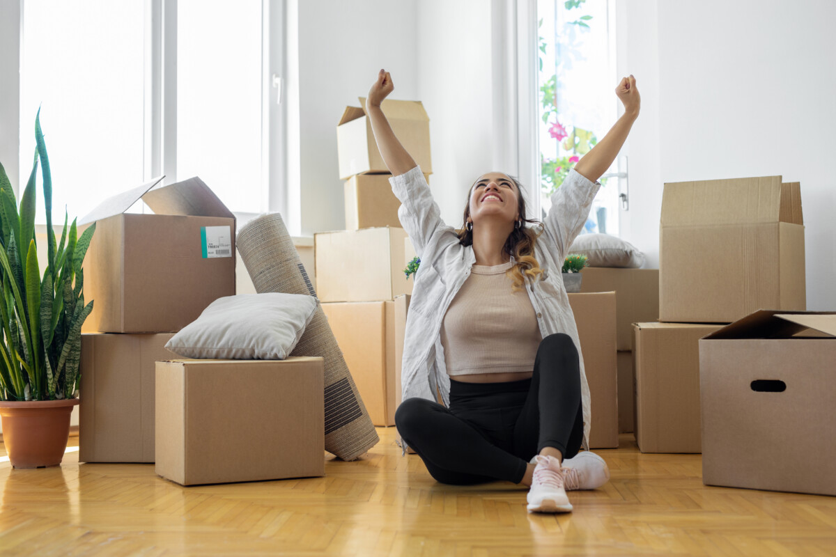 Young woman sitting on floor in new apartment with boxes and raising arms in joy _ getty