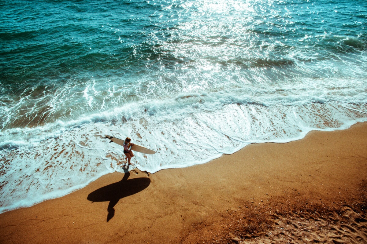 Aerial view photo of a surfer girl running with her surfboard and catching the waves
